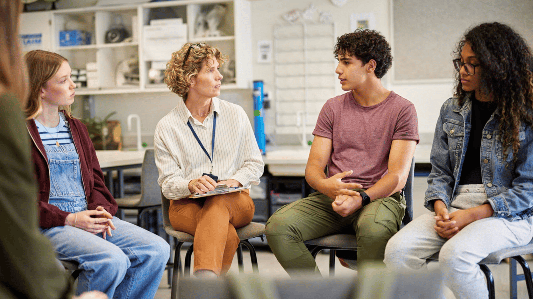 A female educator sitting in a circle with middle and high school students in a well-lit classroom, leading a collaborative mental health awareness and help-seeking discussion.