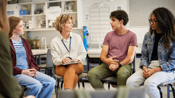 A female educator sitting in a circle with middle and high school students in a well-lit classroom, leading a collaborative mental health awareness and help-seeking discussion.