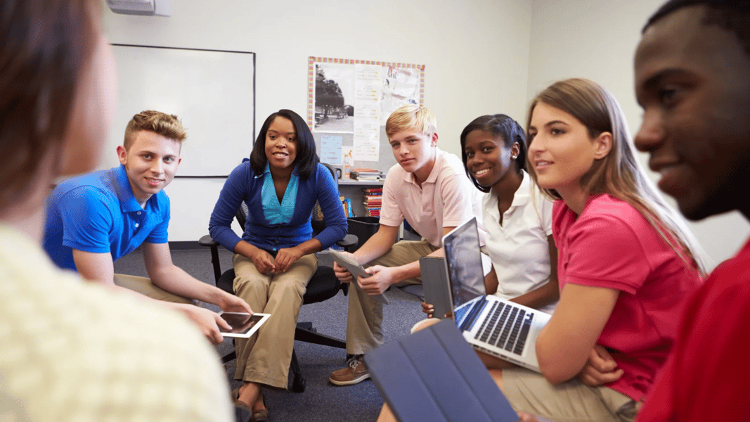 A school counselor and a diverse group of middle and high school students sitting in a circle discussing learning activities using laptops and tablets in a classroom.